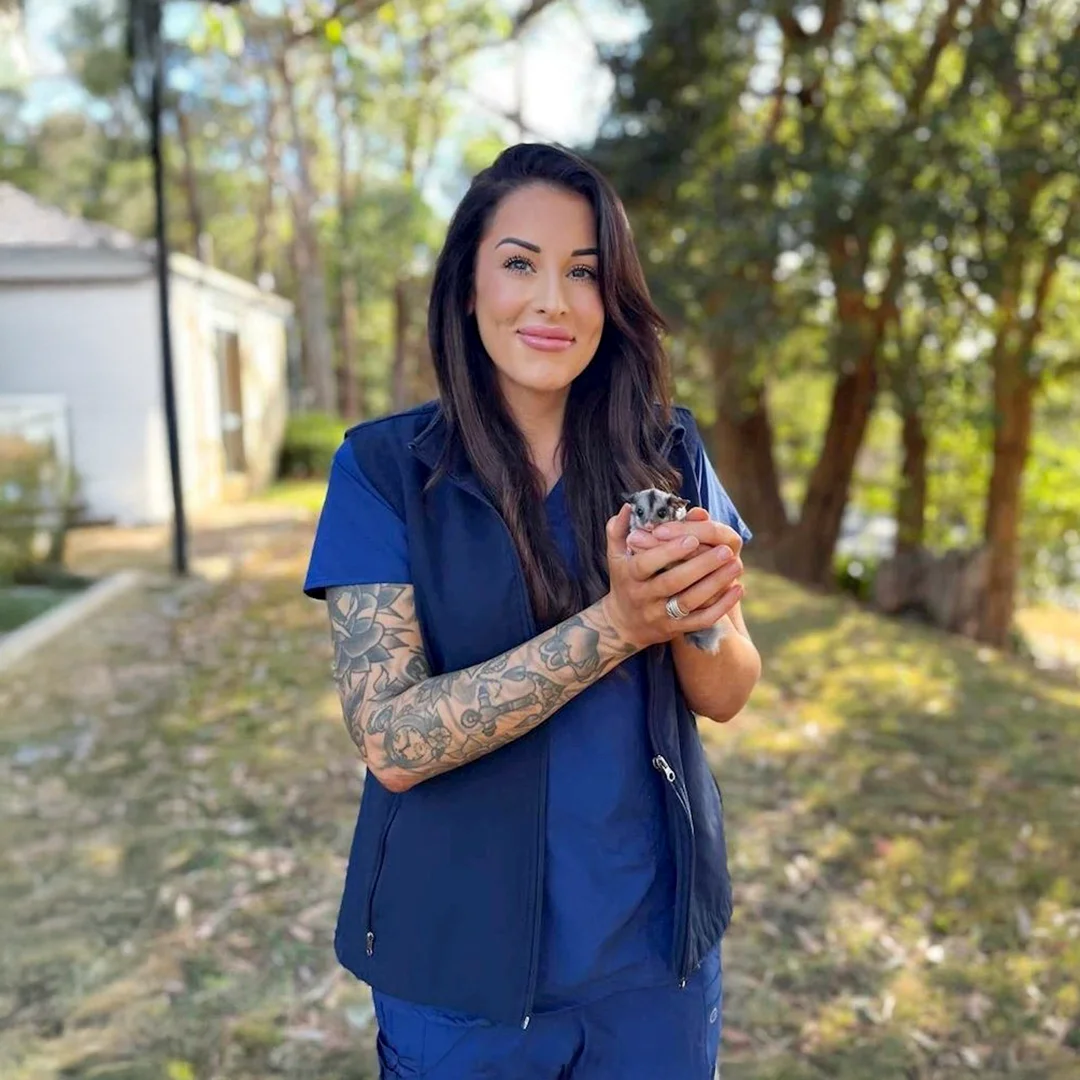 Woman in navy scrubs with tattooed arms holding a sugar glider while standing outside in a sunny, tree-filled area