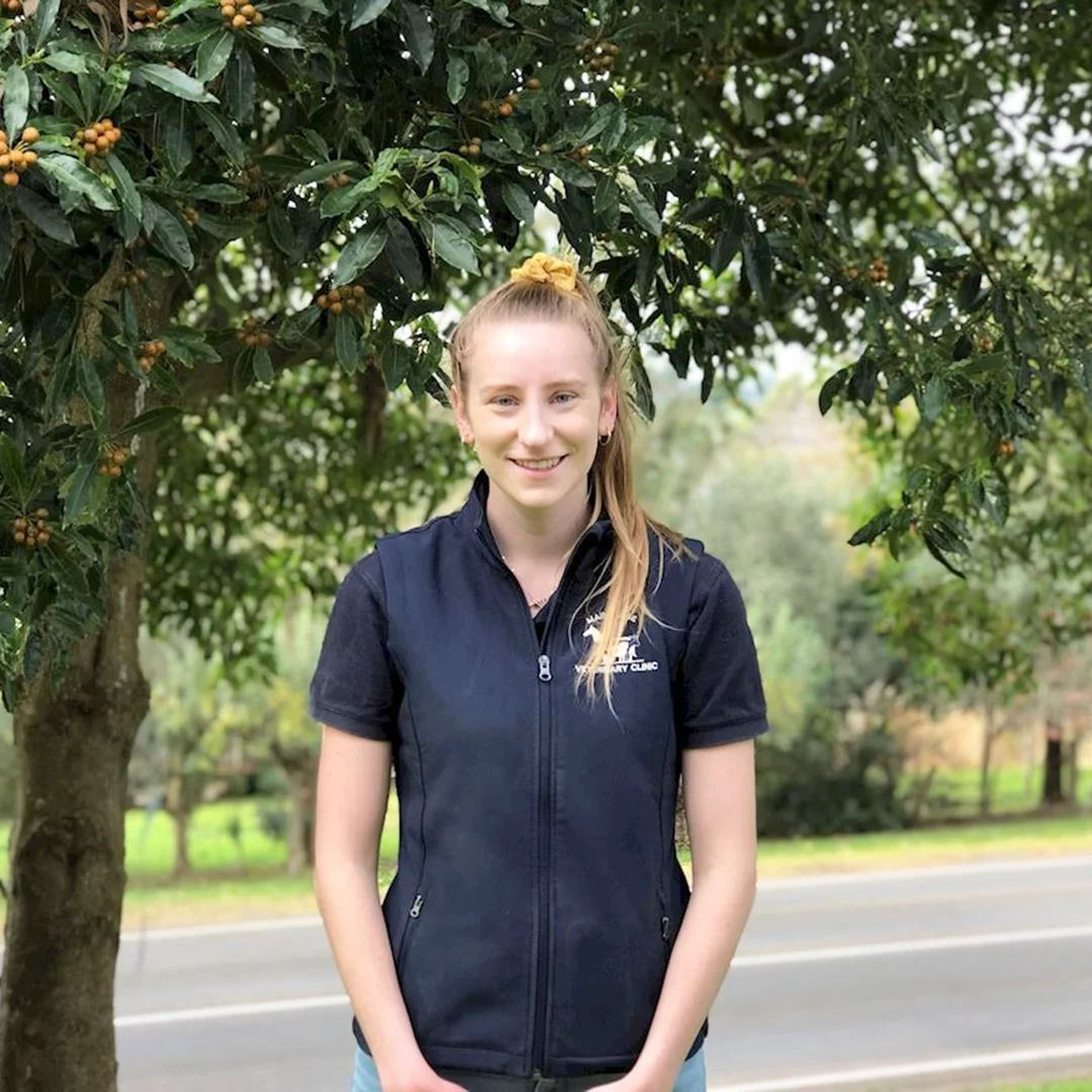 Woman wearing a navy Main Ridge Veterinary Clinic vest standing under a fruiting tree near a country road