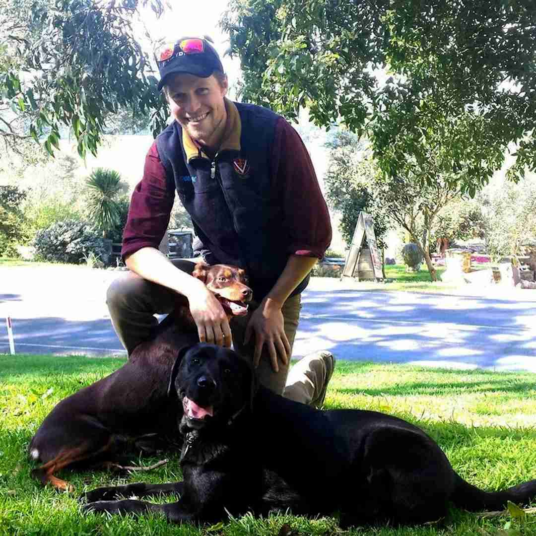 Dr Ben McCubbin with Two Dogs Outdoors Man kneeling on grass smiling with a Doberman and a black Labrador in front of a sunny rural road and garden setting