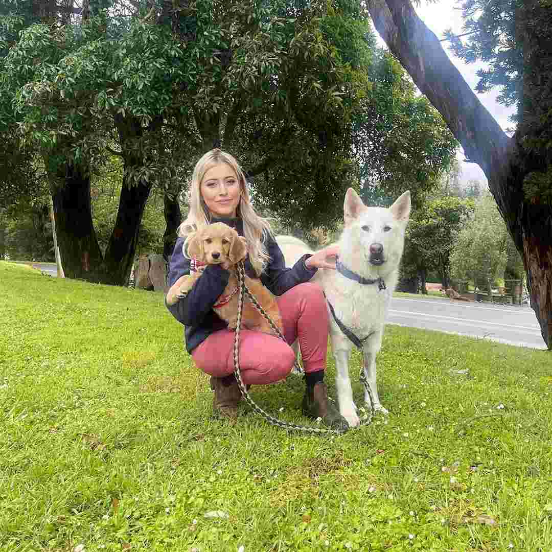 Dr Jessica Beattie with Two Dogs Outdoors Woman crouching on grass holding a tan puppy and petting a white dog on lead in a lush, tree-lined rural setting