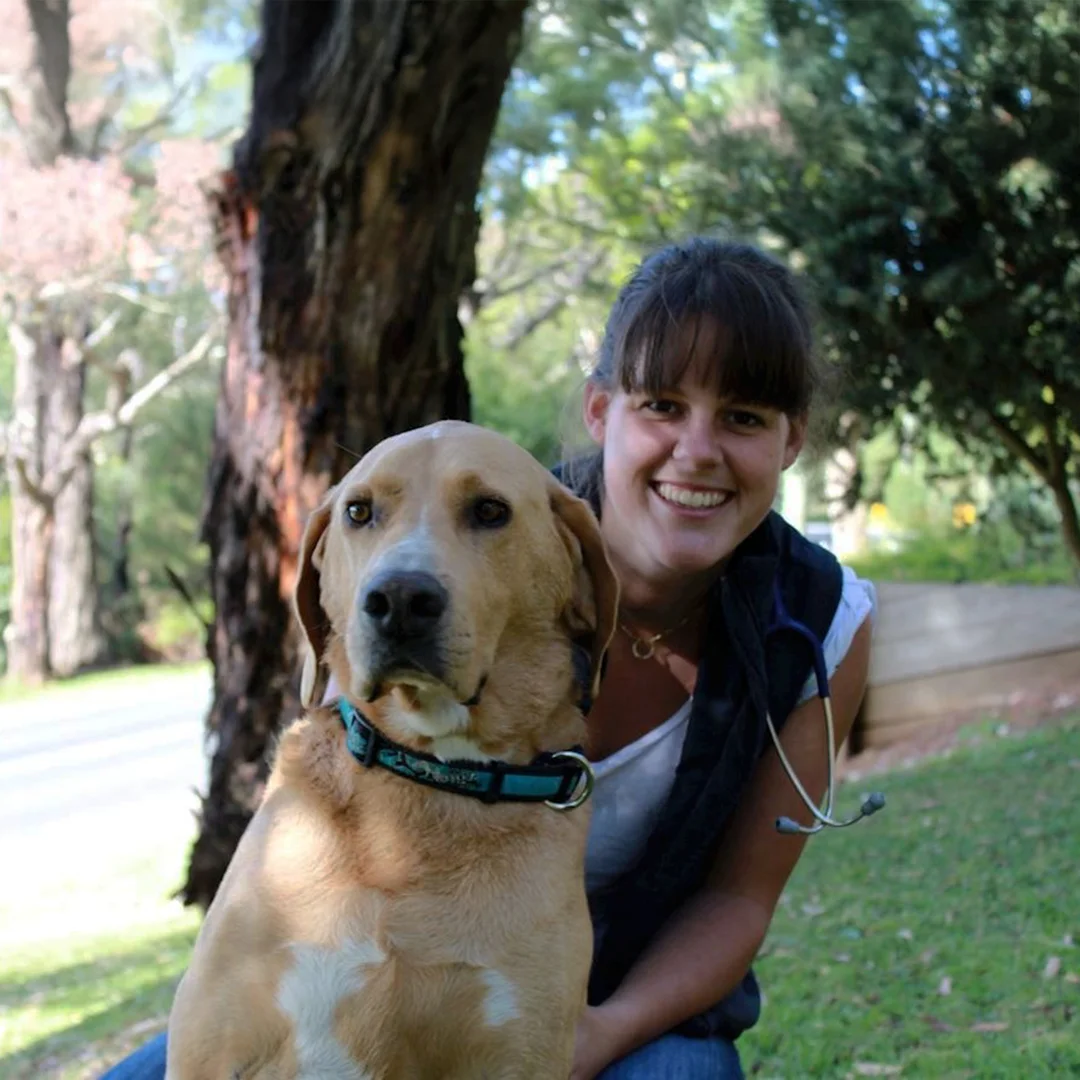 Woman smiling next to a large tan dog wearing a teal collar in a shaded outdoor area with trees and grass