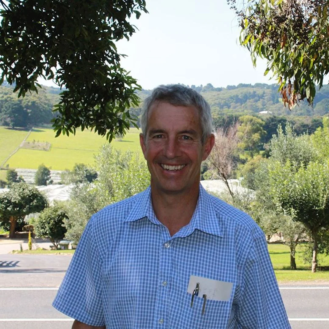 Man smiling in a blue checkered shirt with pen and paper in pocket, standing in front of a tree-lined road and green rolling hills